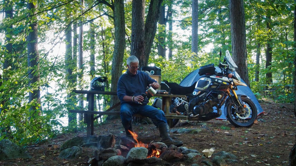 man sitting at picnic table in front of fire pouring coffee. motorcycle and tent in background. view of lake.
