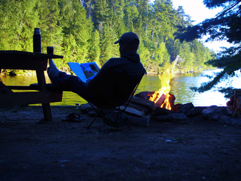 a man sitting by a campfire at the waters edge reading a magazine at the best campground in ontario